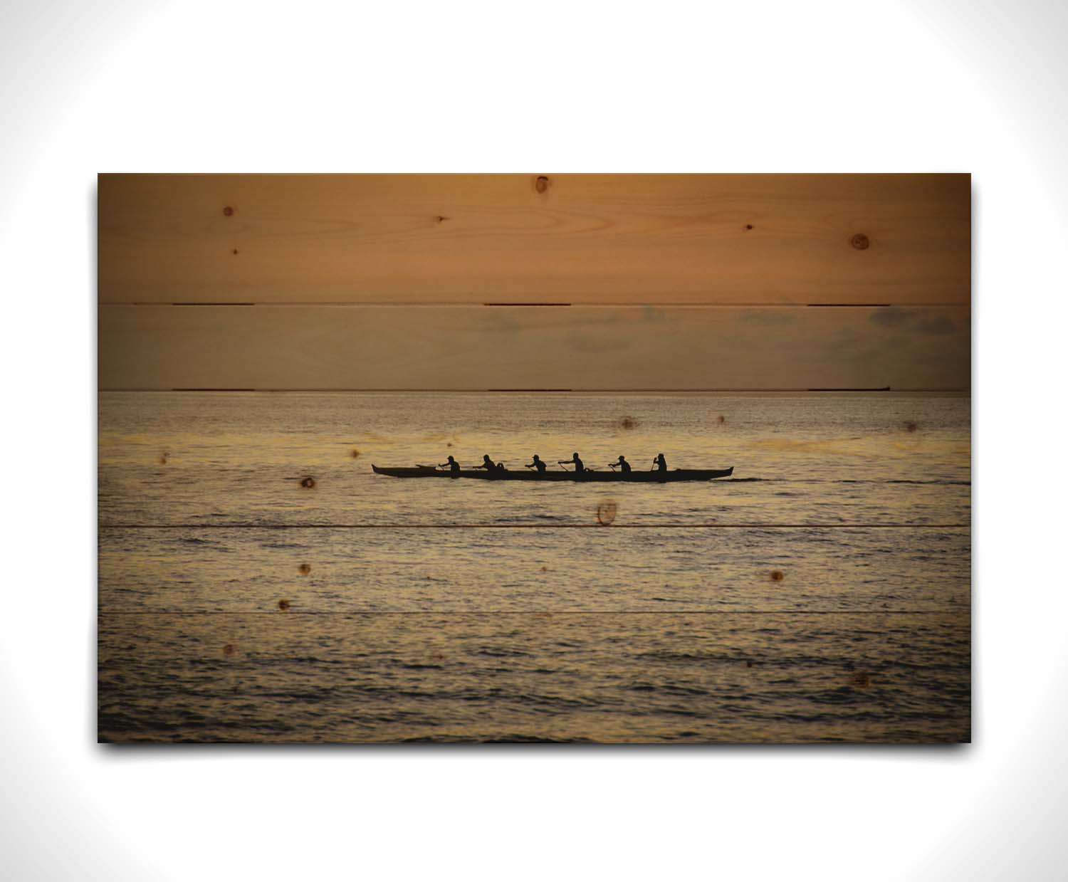 A photo of an outrigger canoe at sea, silhouetted against an orange sky. Printed on a wood pallet.