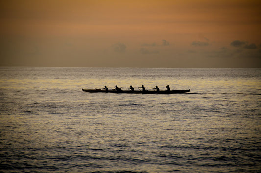 A photo of an outrigger canoe at sea, silhouetted against an orange sky.