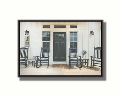 A photo of the door and front porch of a white house, with four black rocking chairs and a black door. Printed on canvas in a float frame.