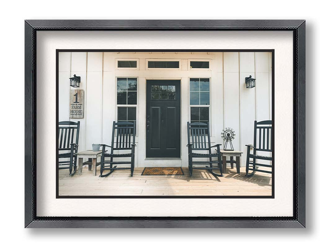 A photo of the door and front porch of a white house, with four black rocking chairs and a black door. Printed on paper, matted, and framed.