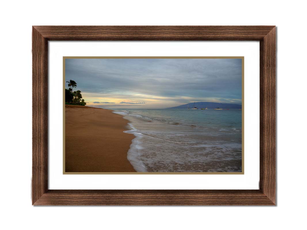 A photo of a cloudy sunrise on the Kaʻanapali shore, looking out to sea. Sailboats can be seen out on the water. Printed on paper, matted, and framed.