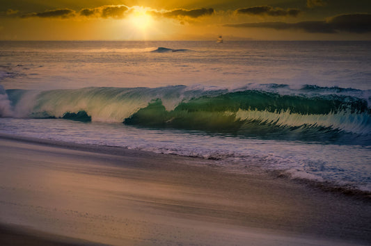 A photo of a calm sea wave rolling onto the beach on Maui at sunset. A boat can be seen on the water in the distance.