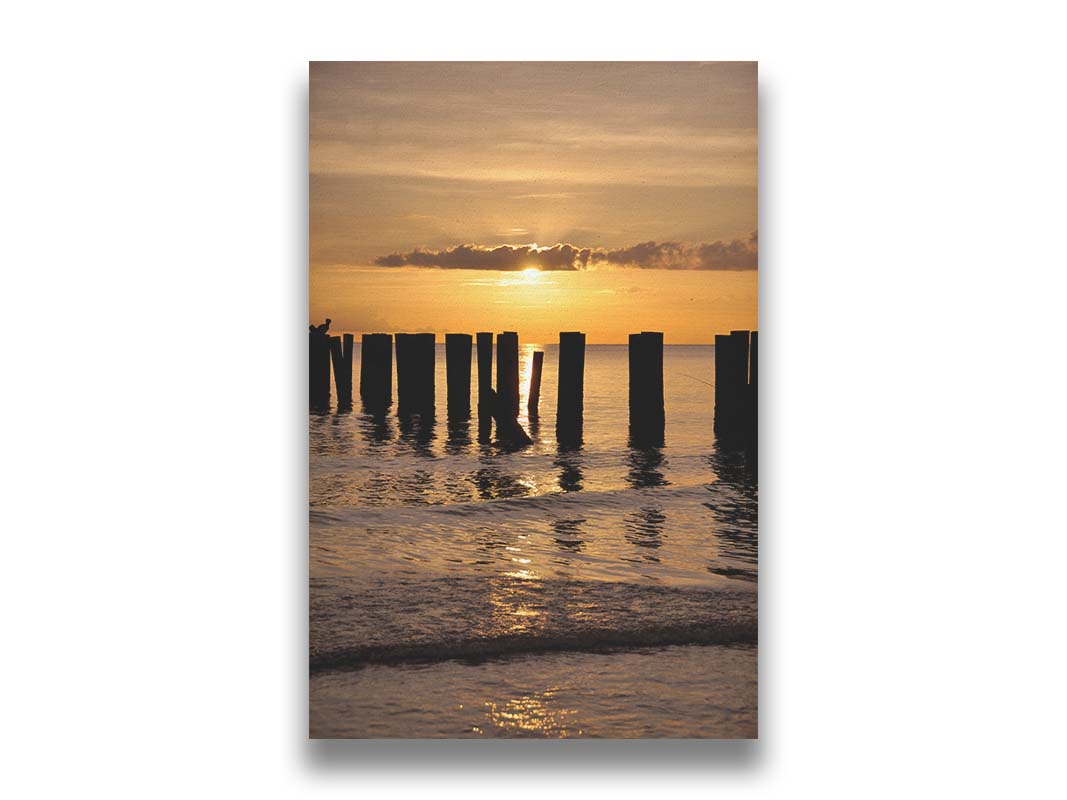 A photo of the remains of the Naples Beach pier in Florida. The sun is low on the horison and silhouettes the pillars in the sea. Printed on canvas.