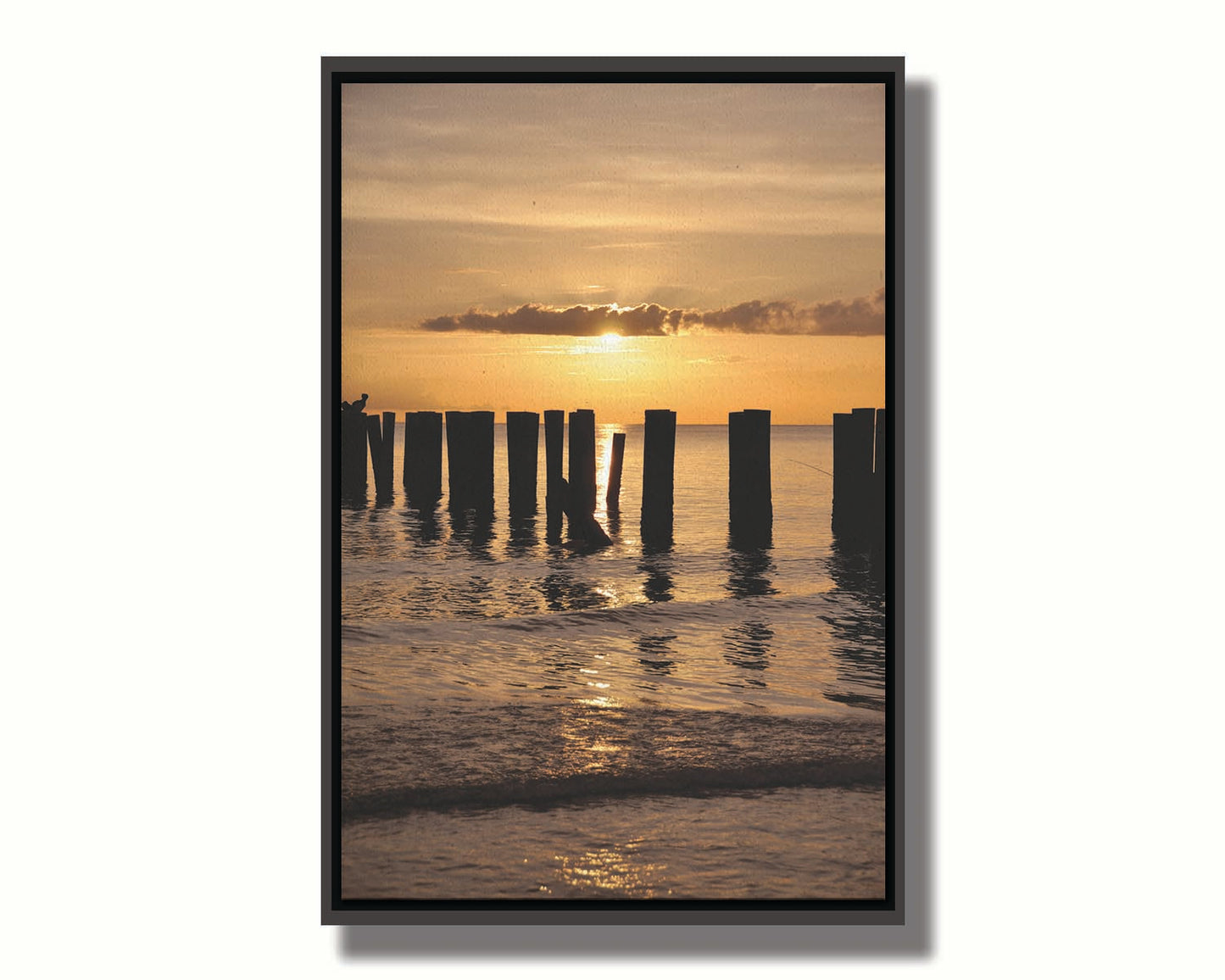 A photo of the remains of the Naples Beach pier in Florida. The sun is low on the horison and silhouettes the pillars in the sea. Printed on canvas in a float frame.