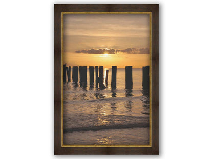 A photo of the remains of the Naples Beach pier in Florida. The sun is low on the horison and silhouettes the pillars in the sea. Printed on canvas and framed.