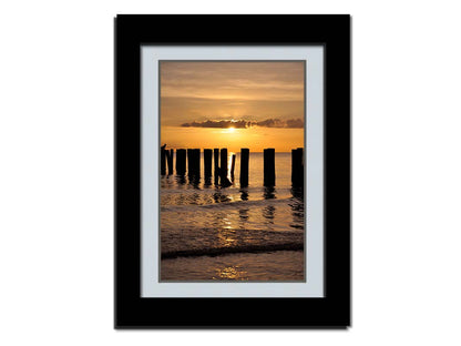 A photo of the remains of the Naples Beach pier in Florida. The sun is low on the horison and silhouettes the pillars in the sea. Printed on paper, matted, and framed.