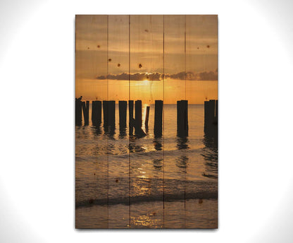 A photo of the remains of the Naples Beach pier in Florida. The sun is low on the horison and silhouettes the pillars in the sea. Printed on a wood pallet.