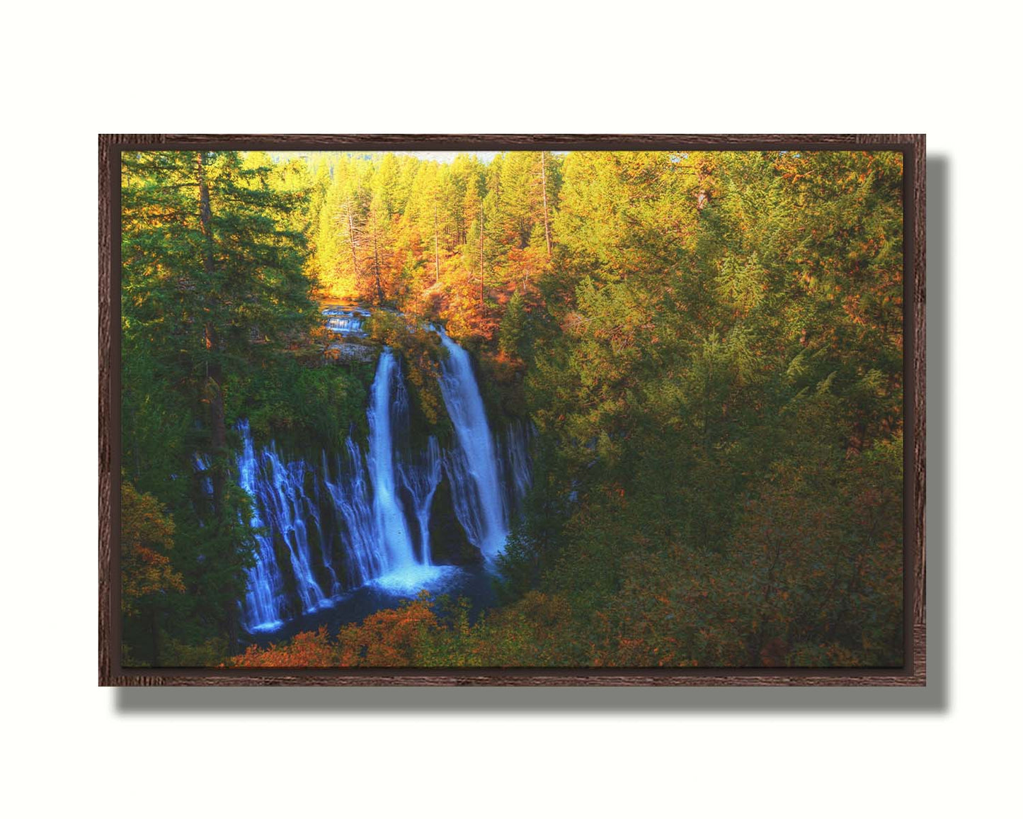 A photograph of Burney Falls in California. Taken from above and away from the falls, the photo gives a full view of the water pouring down the cliff face. The surrounding forest is beginning to turn orange for autumn. Printed on canvas in a float frame.