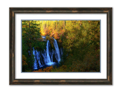 A photograph of Burney Falls in California. Taken from above and away from the falls, the photo gives a full view of the water pouring down the cliff face. The surrounding forest is beginning to turn orange for autumn. Printed on paper, matted, and framed.