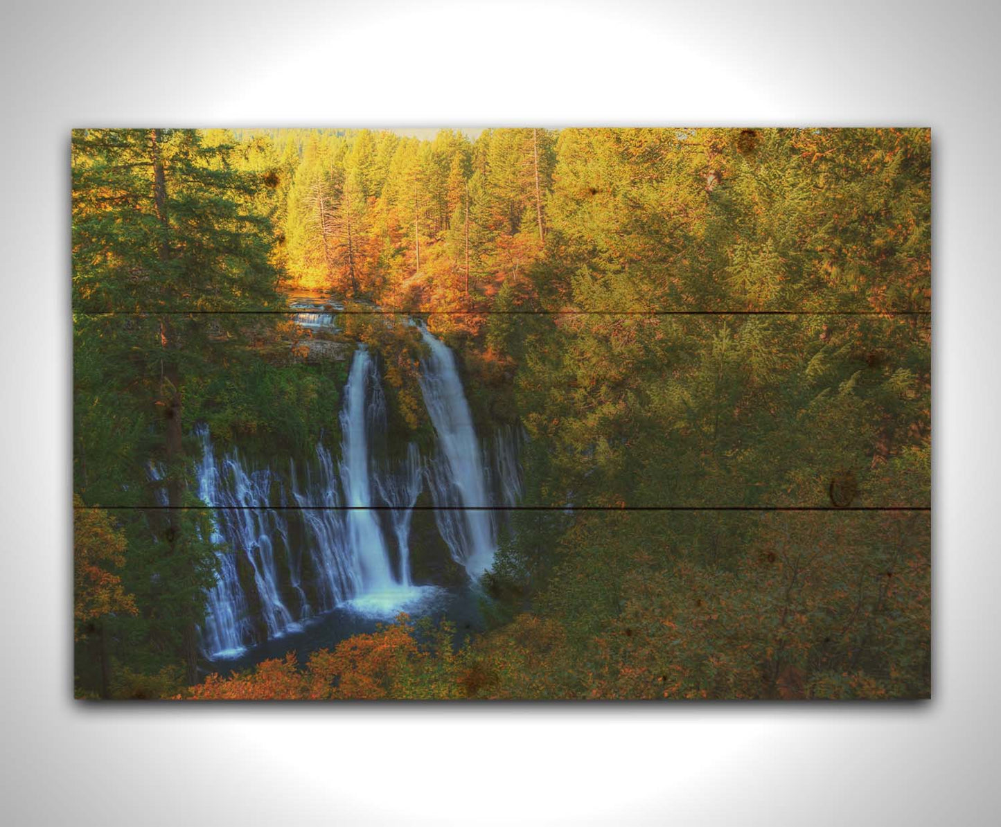 A photograph of Burney Falls in California. Taken from above and away from the falls, the photo gives a full view of the water pouring down the cliff face. The surrounding forest is beginning to turn orange for autumn. Printed on a wood pallet.