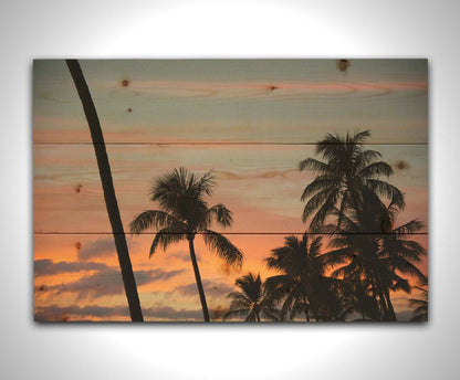 A photograph of Hawaiʻian palm trees, sihouetted against a sunset sky in orange, pink, and blue. Printed on a wood pallet.