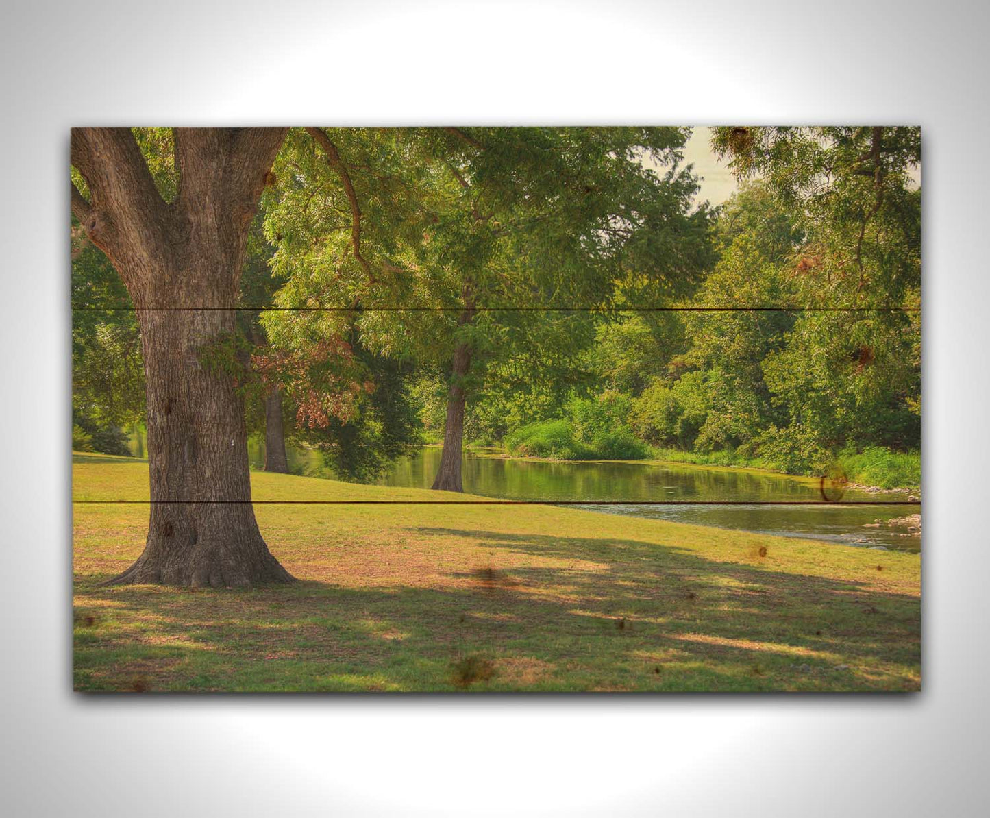 A photograph of a portion of the Comal River through New Braunfels, Texas. Short trimmed grass and park trees line the bank. Printed on a wood pallet.