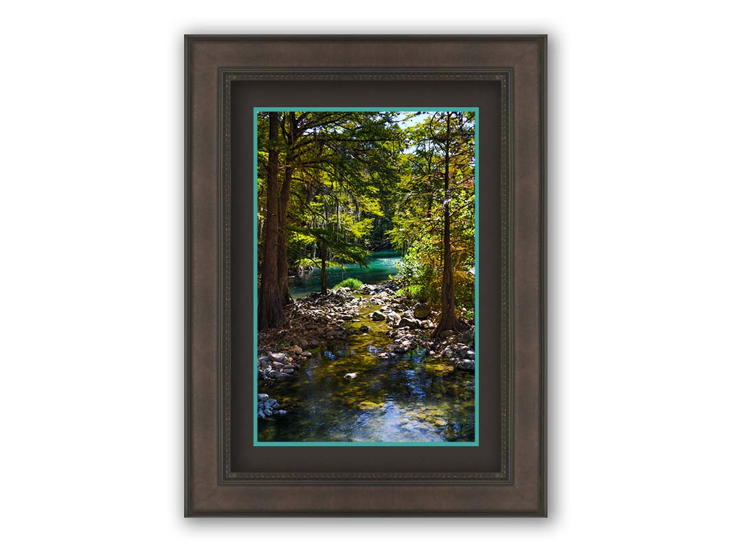 A photograph of the Guadalupe River in Gruene, Texas. Trees and foliage thrive around the water source, creating a lush green scene. The river reflects a vibrant turquoise behind the trees. Printed on paper, matted, and framed.