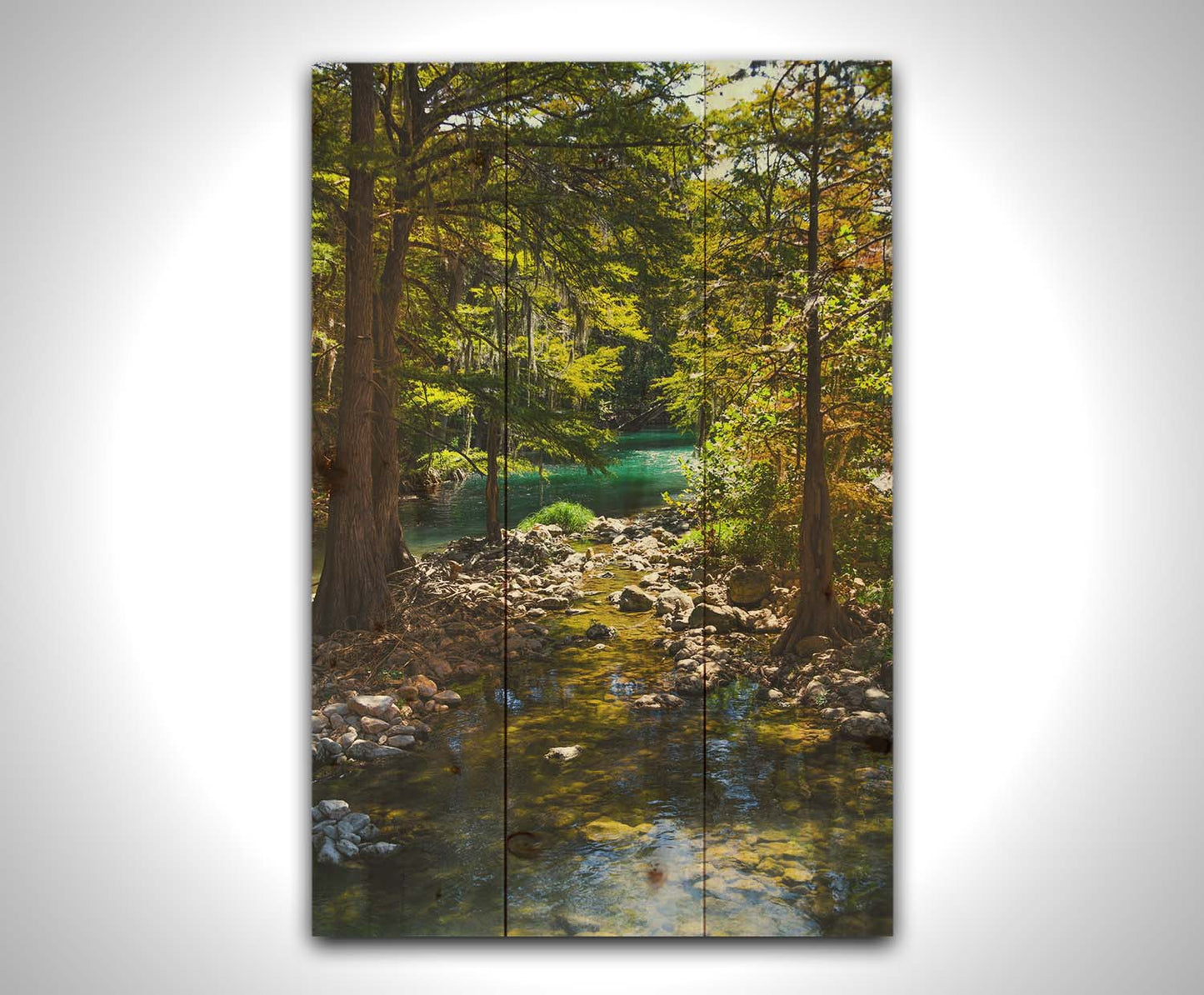 A photograph of the Guadalupe River in Gruene, Texas. Trees and foliage thrive around the water source, creating a lush green scene. The river reflects a vibrant turquoise behind the trees. Printed on a wood pallet.