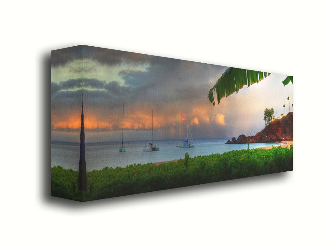 A photograph of Ka'anapali in Maui, Hawai'i. Lush green plants grow on the beach, and sailboats float out on the water. A rainbow pokes down through the clouds before the sunset. Printed on canvas.