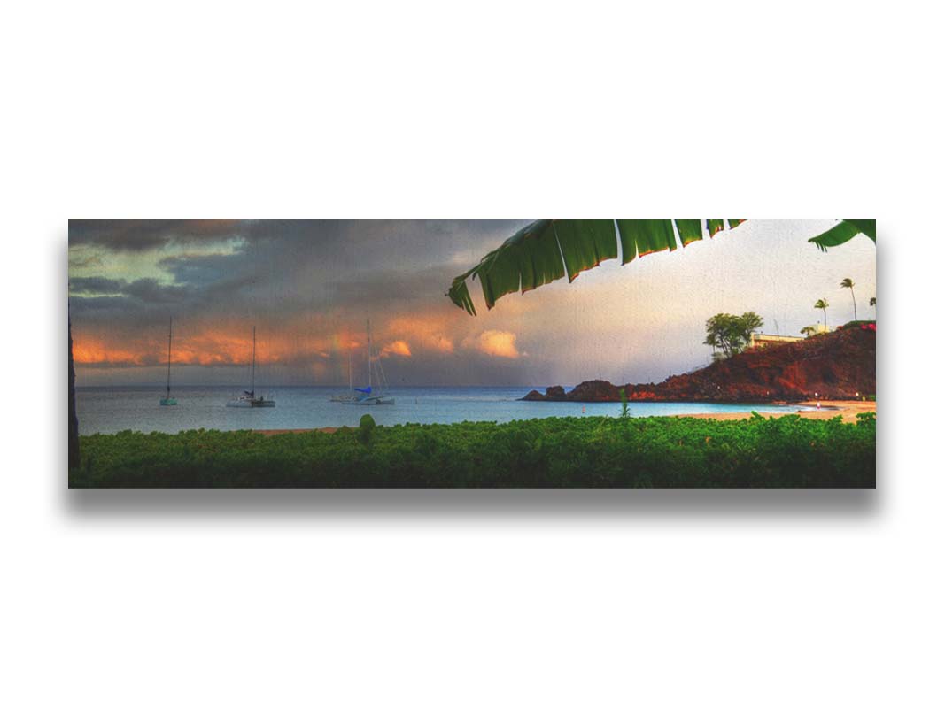 A photograph of Ka'anapali in Maui, Hawai'i. Lush green plants grow on the beach, and sailboats float out on the water. A rainbow pokes down through the clouds before the sunset. Printed on canvas.