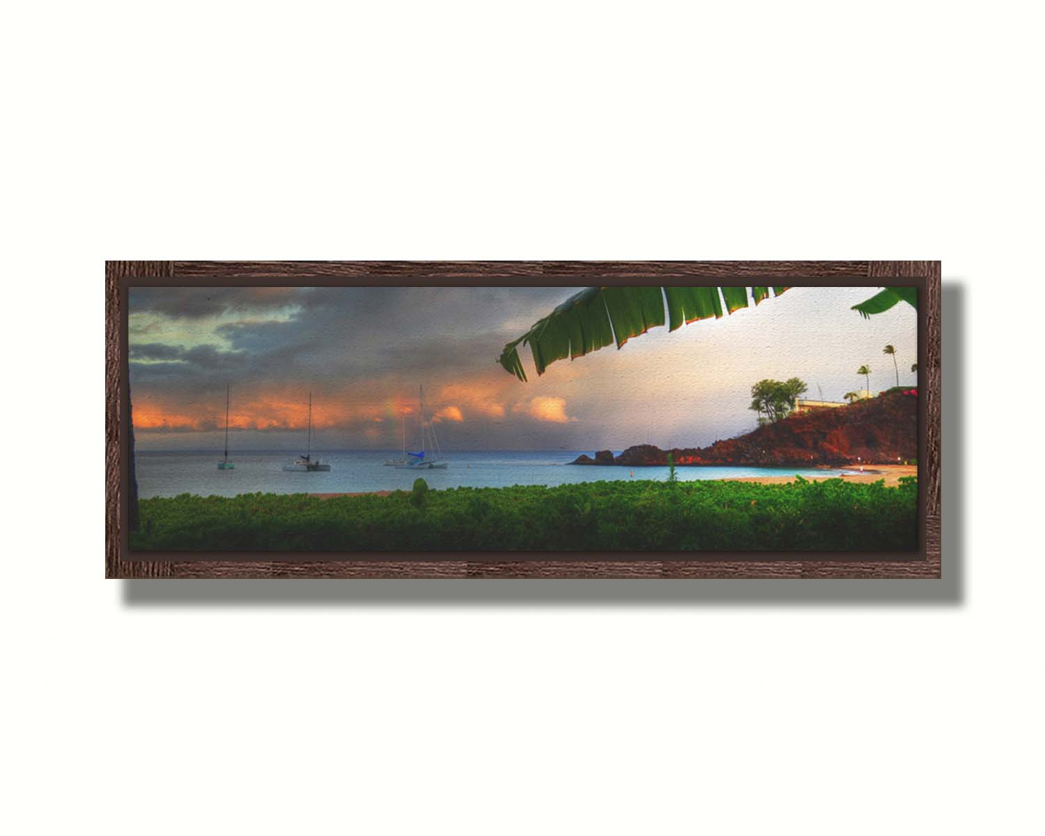 A photograph of Ka'anapali in Maui, Hawai'i. Lush green plants grow on the beach, and sailboats float out on the water. A rainbow pokes down through the clouds before the sunset. Printed on canvas in a float frame.