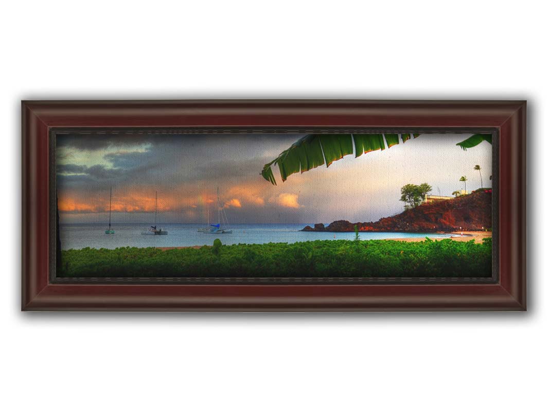 A photograph of Ka'anapali in Maui, Hawai'i. Lush green plants grow on the beach, and sailboats float out on the water. A rainbow pokes down through the clouds before the sunset. Printed on canvas and framed.