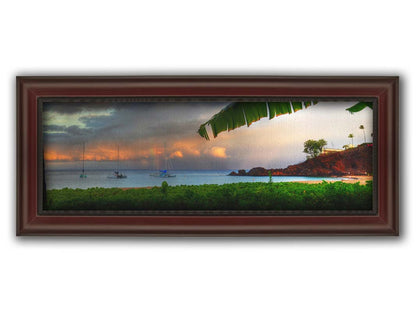 A photograph of Ka'anapali in Maui, Hawai'i. Lush green plants grow on the beach, and sailboats float out on the water. A rainbow pokes down through the clouds before the sunset. Printed on canvas and framed.