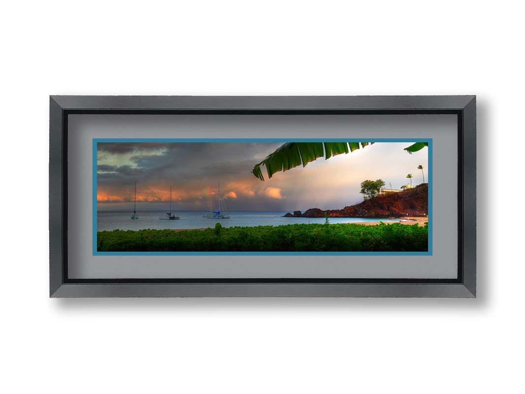 A photograph of Ka'anapali in Maui, Hawai'i. Lush green plants grow on the beach, and sailboats float out on the water. A rainbow pokes down through the clouds before the sunset. Printed on paper, matted, and framed.