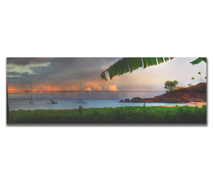 A photograph of Ka'anapali in Maui, Hawai'i. Lush green plants grow on the beach, and sailboats float out on the water. A rainbow pokes down through the clouds before the sunset. Printed on a box board.