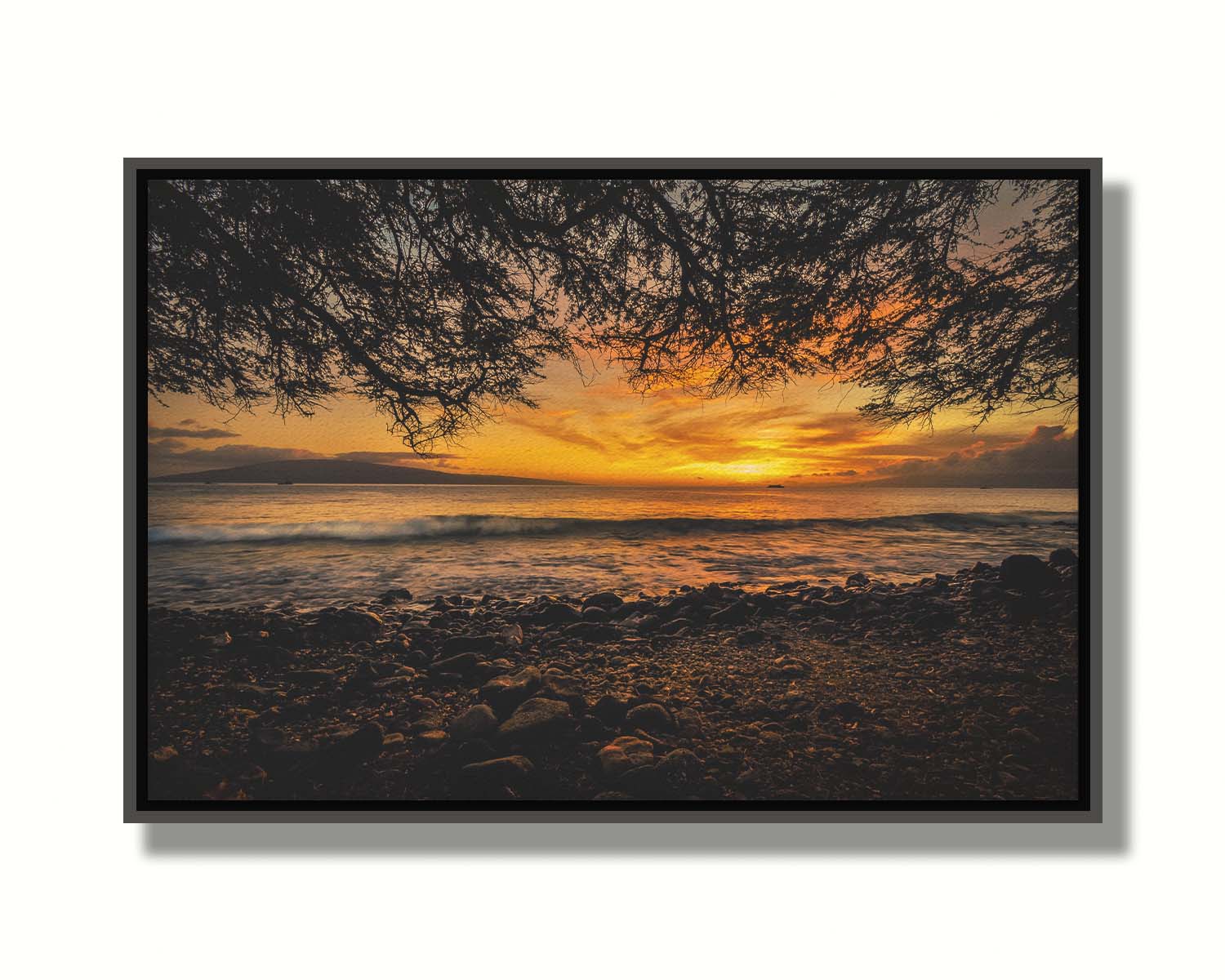 A photograph of the sunset over the ocean from Lāhainā in Maui, Hawaiʻi on a rocky beach, from underneath a tree. Printed on canvas in a float frame.