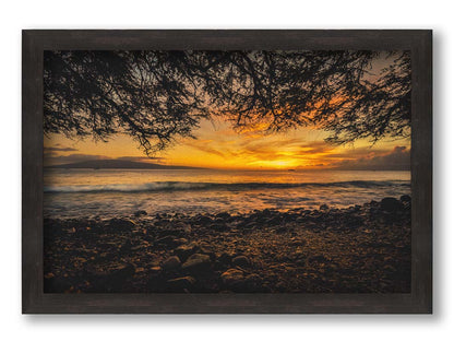 A photograph of the sunset over the ocean from Lāhainā in Maui, Hawaiʻi on a rocky beach, from underneath a tree. Printed on canvas and framed.