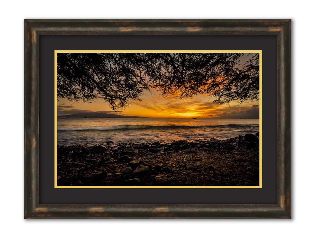 A photograph of the sunset over the ocean from Lāhainā in Maui, Hawaiʻi on a rocky beach, from underneath a tree. Printed on paper, matted, and framed.