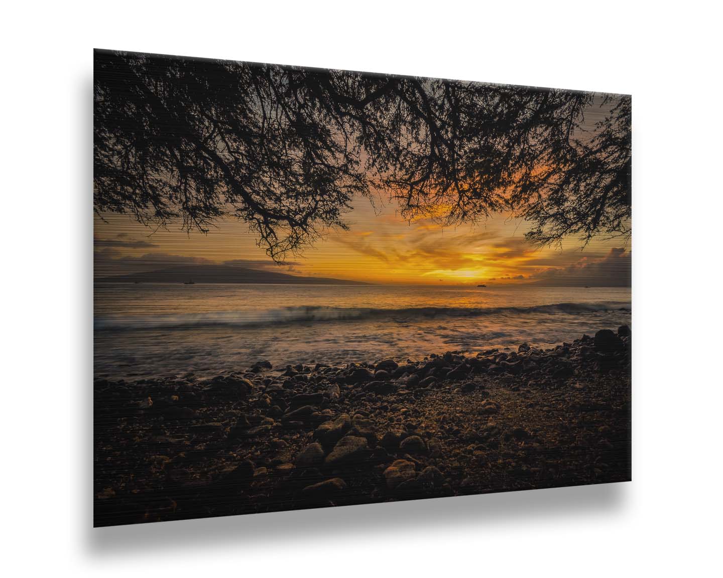 A photograph of the sunset over the ocean from Lāhainā in Maui, Hawaiʻi on a rocky beach, from underneath a tree. Printed on metal.