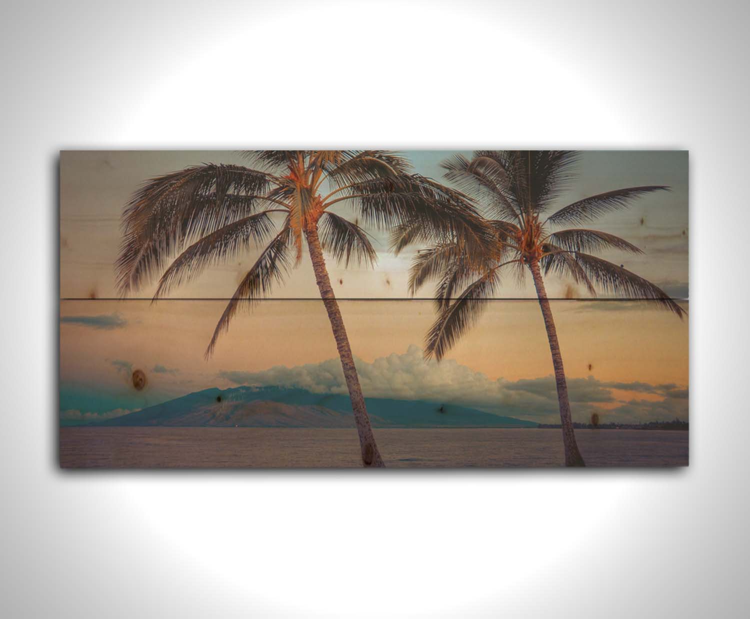 A photo of two palm trees in Maui at sunset, with the sea and cloud-topped mountains in the background. Printed on a wood pallet.