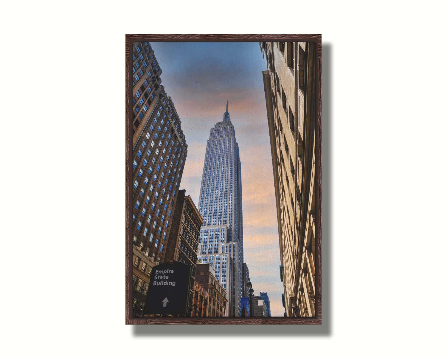 A photoraph looking up at the Empire State Building in New York against a blue sky and orange clouds. Printed on canvas in a float frame.