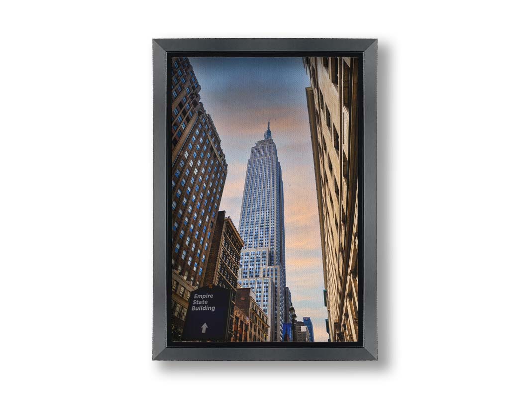 A photoraph looking up at the Empire State Building in New York against a blue sky and orange clouds. Printed on canvas and framed.