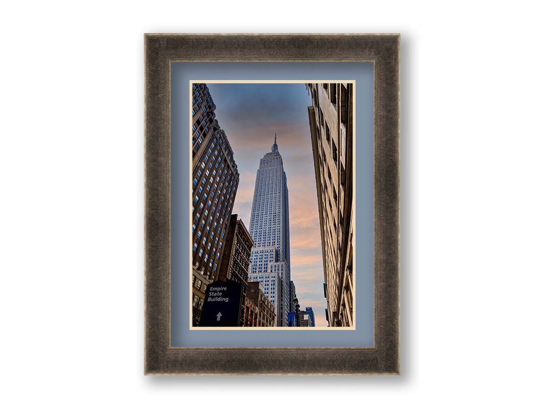 A photoraph looking up at the Empire State Building in New York against a blue sky and orange clouds. Printed on paper, matted, and framed.