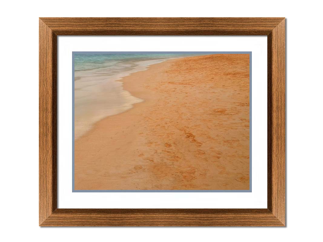 A photograph of beach sand. The warm hues of golden sand are contrasted by the cool ocean water. Printed on paper, matted, and framed.