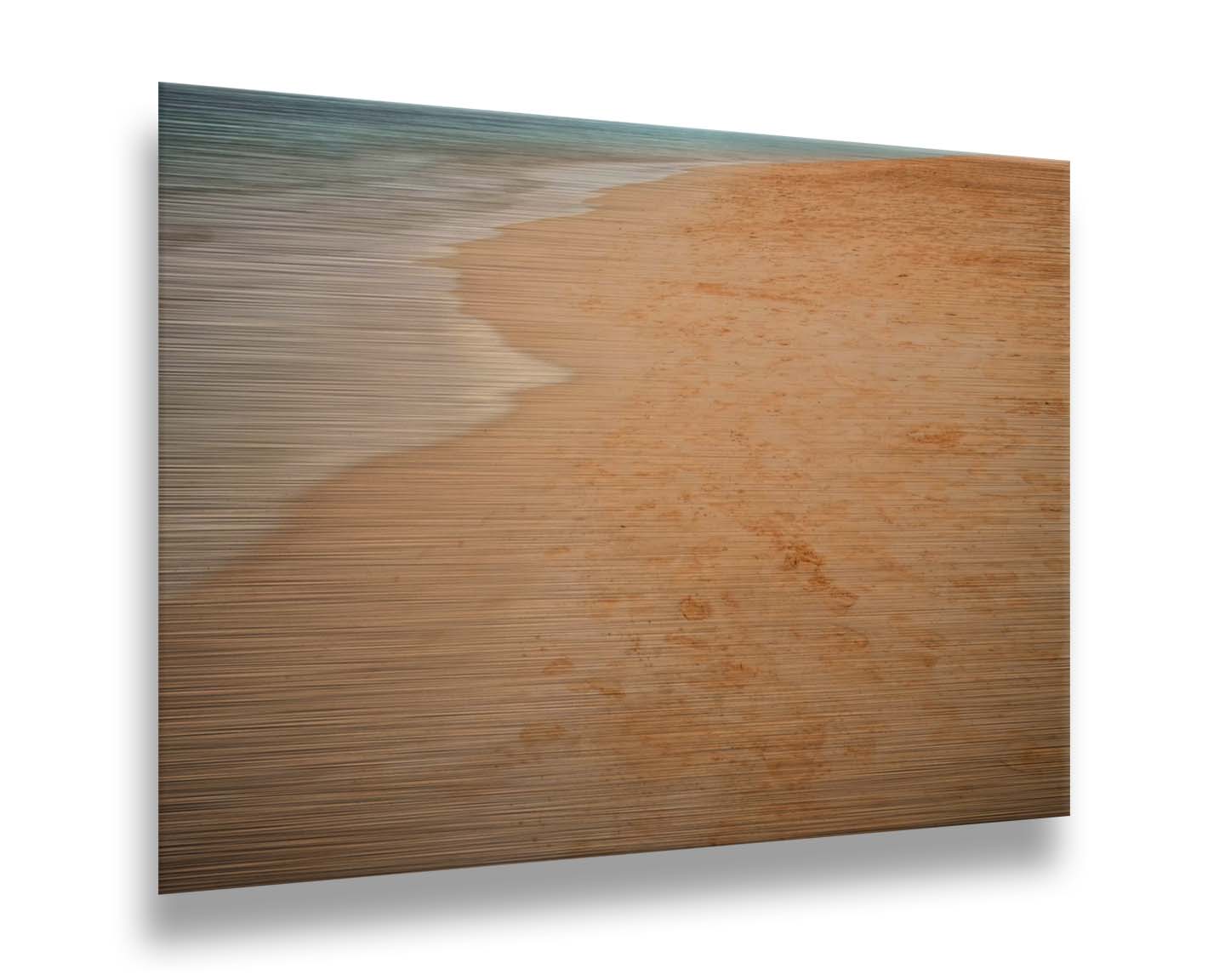 A photograph of beach sand. The warm hues of golden sand are contrasted by the cool ocean water. Printed on metal.