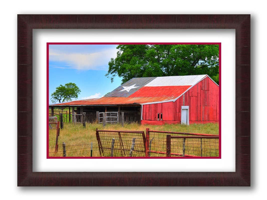 A photograph of a red barn, with a Texas flag painted on the roof. It is surrounded by red ranch fences and grassy fields. Printed on paper, matted, and framed.