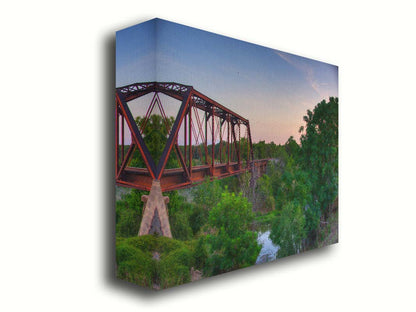 A photograph of a red metal railroad bridge passing over green trees and a river. The low sun turns the blue sky pink. Printed on canvas.