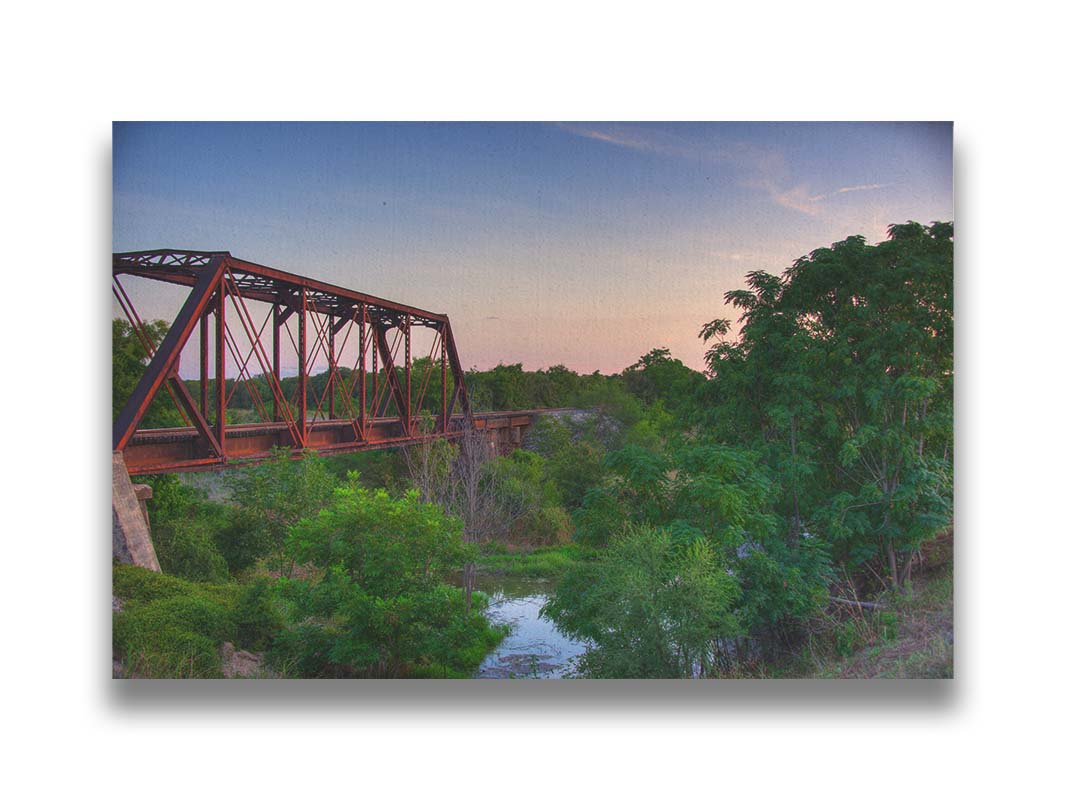 A photograph of a red metal railroad bridge passing over green trees and a river. The low sun turns the blue sky pink. Printed on canvas.