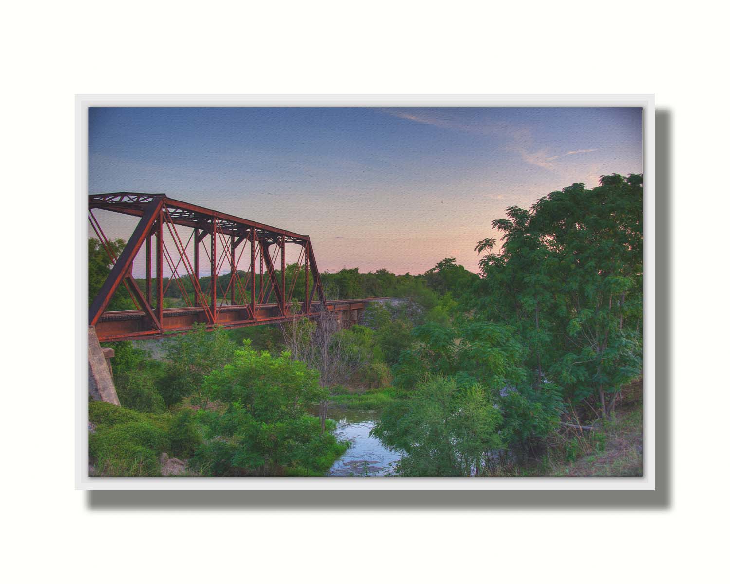 A photograph of a red metal railroad bridge passing over green trees and a river. The low sun turns the blue sky pink. Printed on canvas in a float frame.