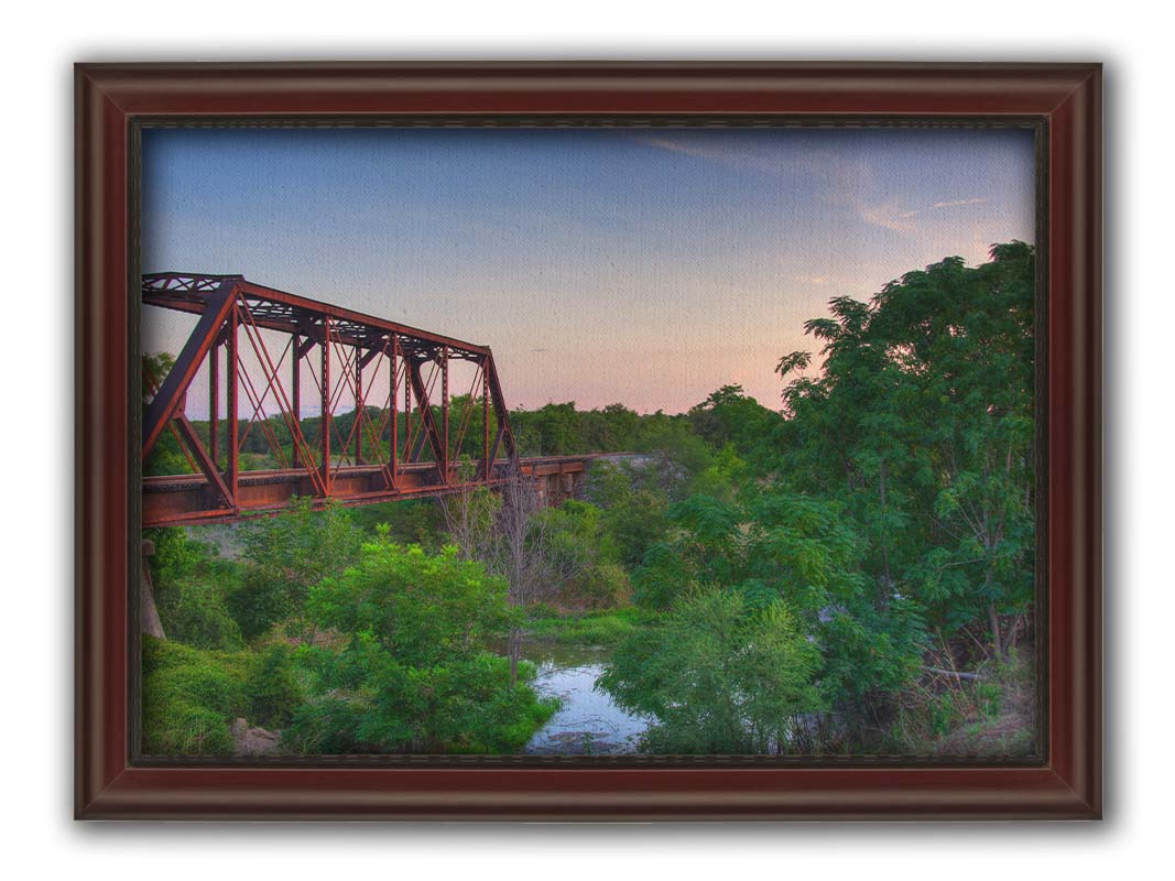 A photograph of a red metal railroad bridge passing over green trees and a river. The low sun turns the blue sky pink. Printed on canvas and framed.