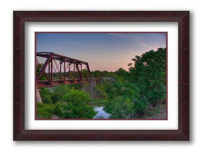 A photograph of a red metal railroad bridge passing over green trees and a river. The low sun turns the blue sky pink. Printed on paper, matted, and framed.