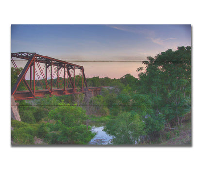 A photograph of a red metal railroad bridge passing over green trees and a river. The low sun turns the blue sky pink. Printed on a box board.