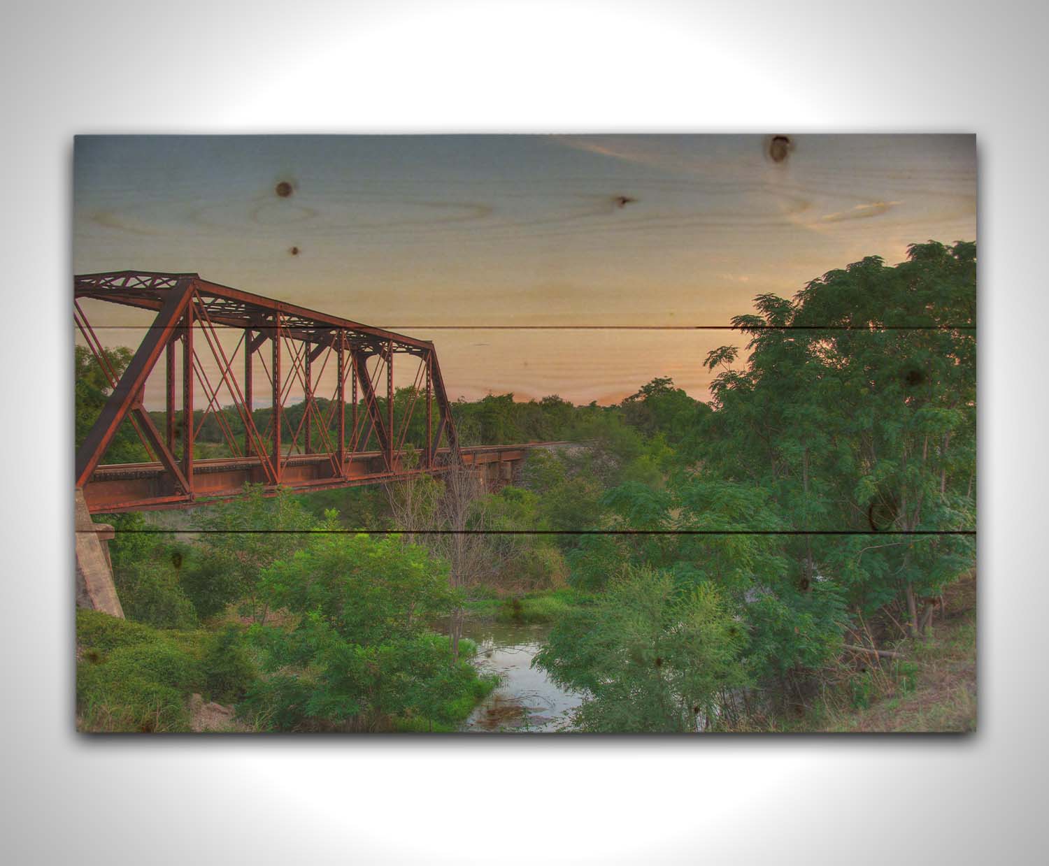 A photograph of a red metal railroad bridge passing over green trees and a river. The low sun turns the blue sky pink. Printed on a wood pallet.