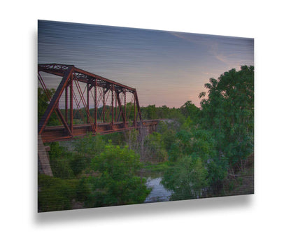 A photograph of a red metal railroad bridge passing over green trees and a river. The low sun turns the blue sky pink. Printed on metal.