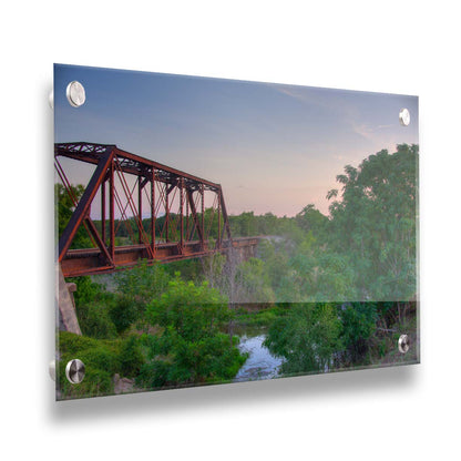 A photograph of a red metal railroad bridge passing over green trees and a river. The low sun turns the blue sky pink. Printed on acrylic.