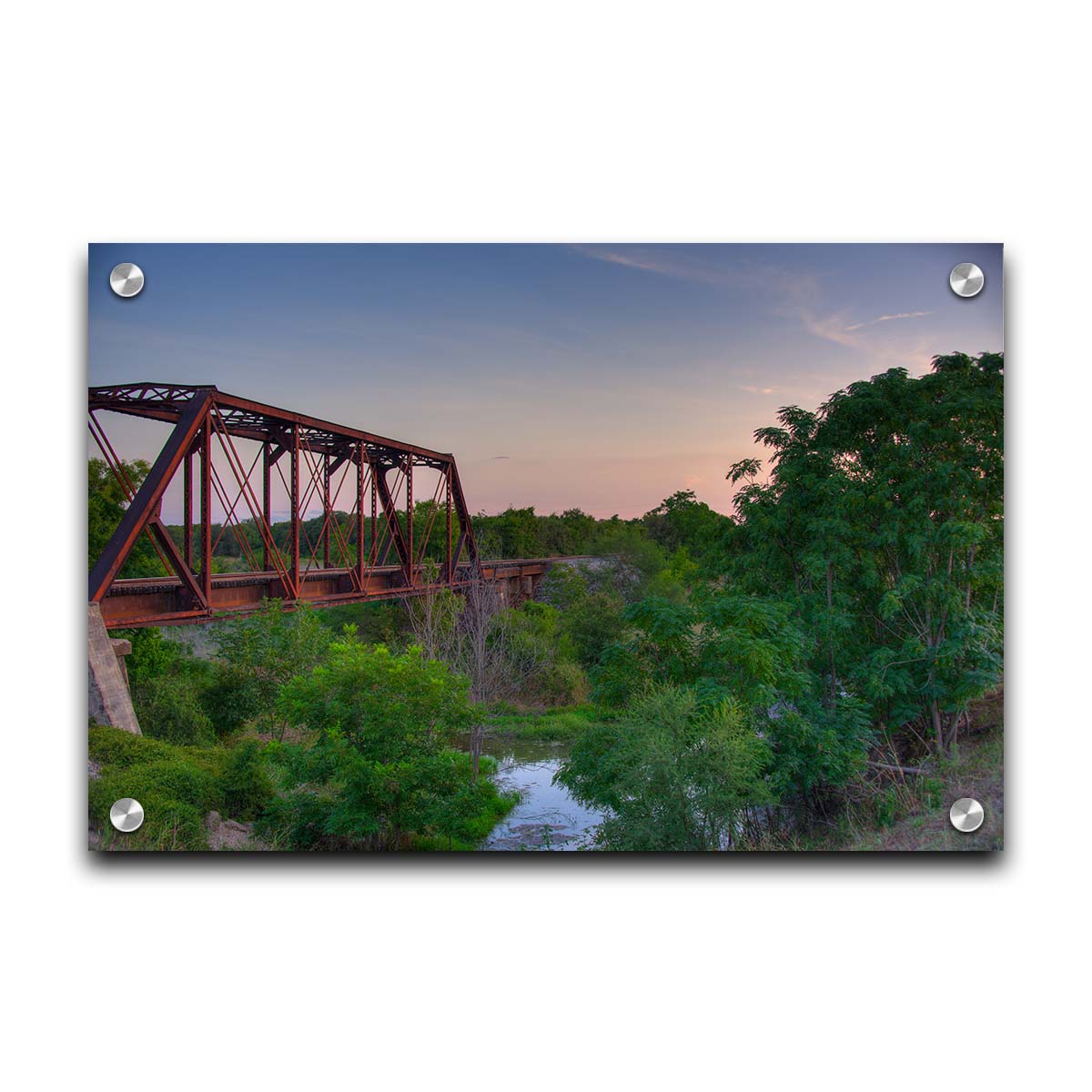 A photograph of a red metal railroad bridge passing over green trees and a river. The low sun turns the blue sky pink. Printed on acrylic.