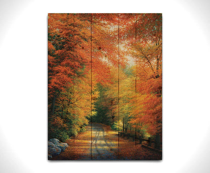 A painting of a road winding through a forest during fall. The red, orange, and yellow leaves shed to the ground, lining the road. Printed on a wood pallet.