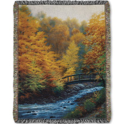 Woven throw blanket designed with a painting of a forest stream, passing under an arched bridge. The trees are changing to yellow and orange for fall.