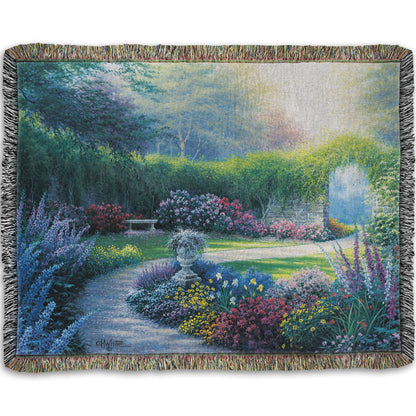 A woven blanket designed with a painting of a serene path through a garden filled with bushes of flowers bathed in a gentle light.