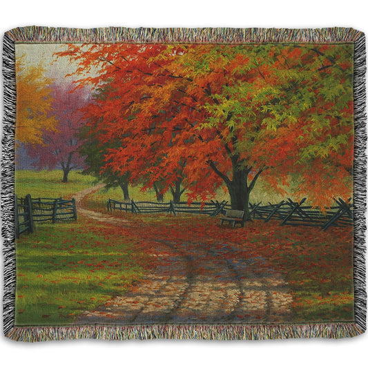 A woven throw blanket designed with a painting of a road winding through a fenced field, surrounded by multicolored fall maple trees.